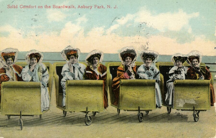 Colored Postcard showing eight women with hats on beach chairs on wheels with heading "Solid comfort on the boardwalk, Asbury Park, N.J."